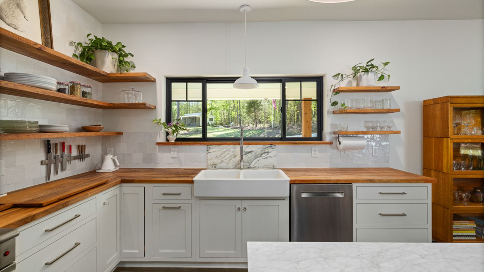 White kitchen with open wood shelves, a farmhouse sink under the window, and empty butcher-block counters in the Nancy Meyers style